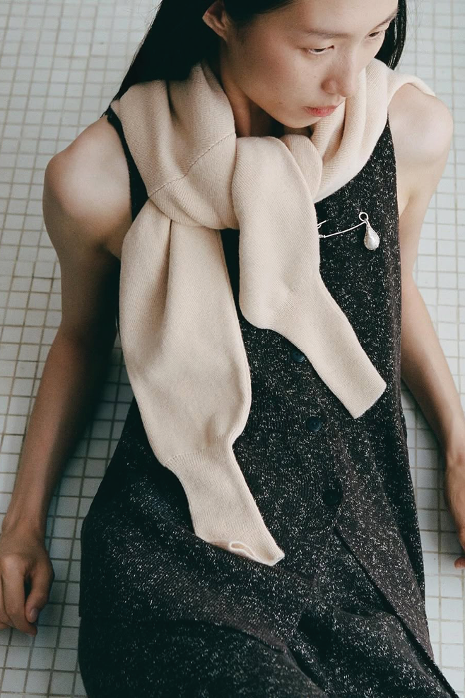 A close-up, high-angle shot of a person sitting down, wearing a dark brown, sleeveless knit dress. A cream or beige knitted sweater is draped around their neck, secured by a large, silver safety pin that holds a teardrop-shaped pearl or pendant.