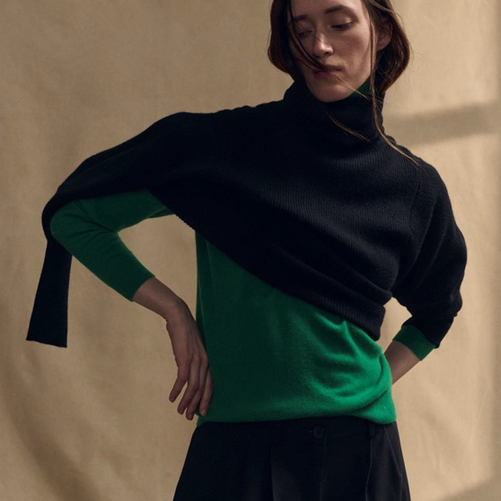 A person wearing a black turtleneck sweater draped over a bright emerald green, long-sleeved turtleneck sweater and a black skirt, posing with hands on hips against a neutral beige background.