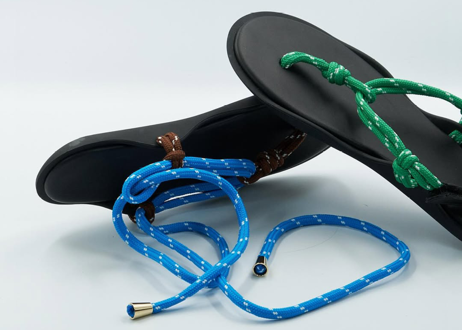 A pair of sandals with black soles are shown on a white background. One sandal features a bright green rope for the toe post and strap, while the other features a brown rope. A loose coil of blue and white dotted rope, seemingly a detachable or alternative strap, lies in the foreground.