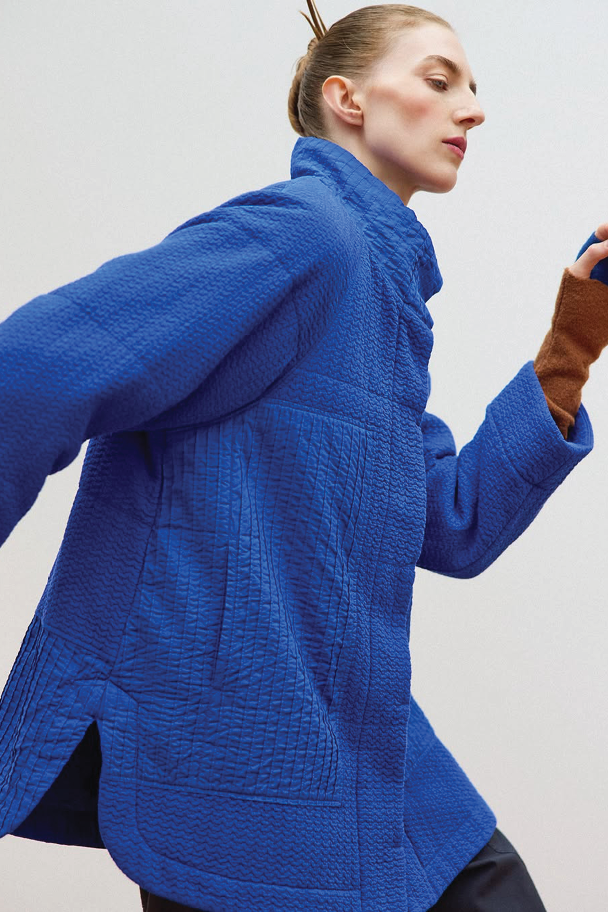 A dynamic shot of a person in motion wearing a vibrant royal blue quilted jacket with intricate vertical stitching and side slits, paired with brown knit fingerless gloves.