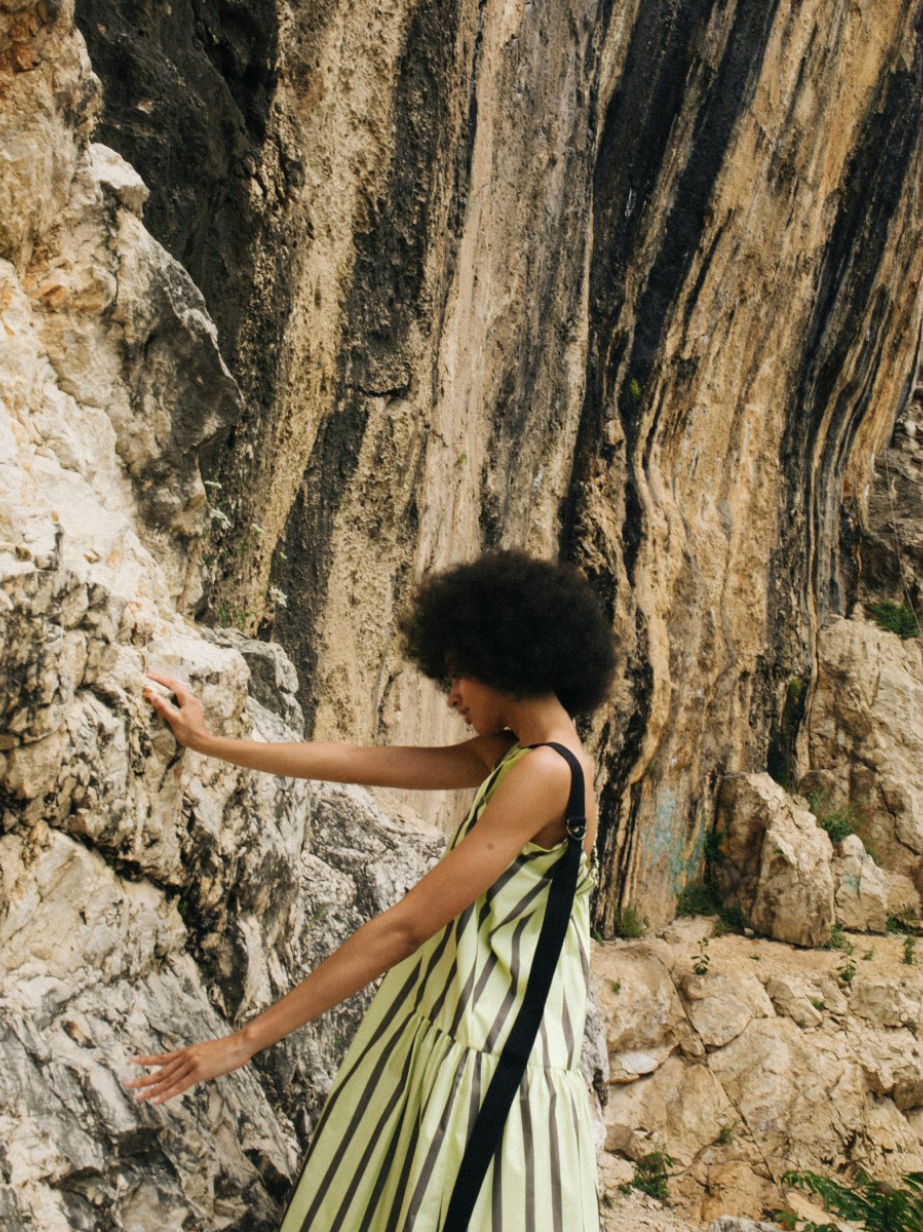A person with a large afro hairstyle standing against a textured rock wall, wearing a pale yellow and dark green vertically striped maxi dress with a tiered skirt and thin black straps.