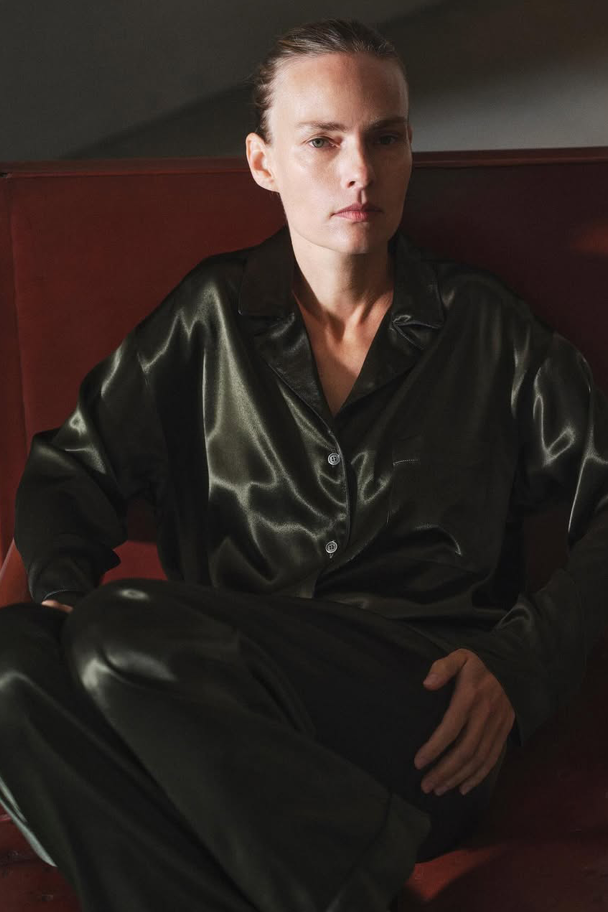 A person seated indoors wearing a luxurious, oversized dark olive green silk or satin pajama-style set, featuring a button-down shirt with a wide collar and matching relaxed-fit trousers.