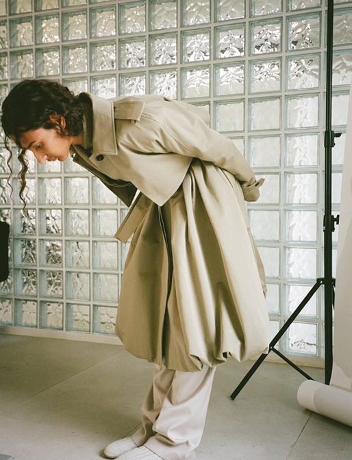 A person with long, wavy dark hair leaning forward in a profile view, showcasing a voluminous, sand-colored trench coat with a structured collar and heavy pleating that creates a bell-like silhouette. They are paired with wide-leg beige trousers and white knit socks, posed against a backdrop of a glass block wall.