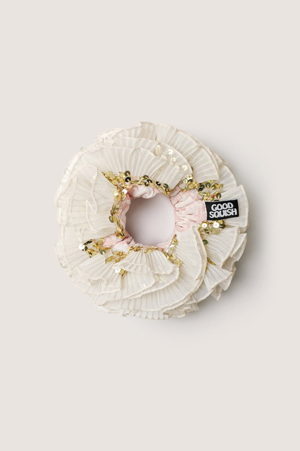 A top-down view of a large, cream-colored ruffled hair accessory featuring gold sequin embellishments and a black "GOOD SQUISH" brand label.