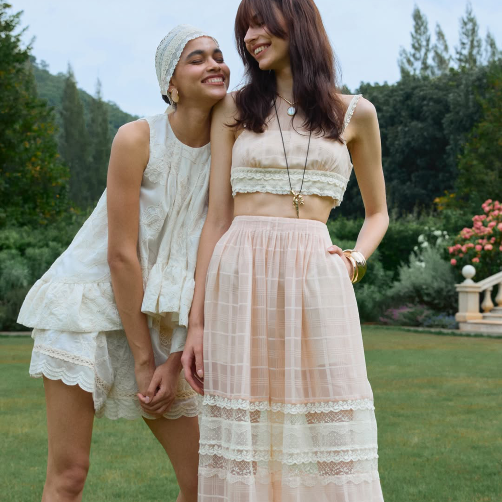 Two people standing outdoors in a garden setting. One wears a white sleeveless lace-trimmed top and shorts set with a matching headwrap; the other wears a peach-colored cropped lace camisole and a matching high-waisted, semi-sheer pleated maxi skirt.