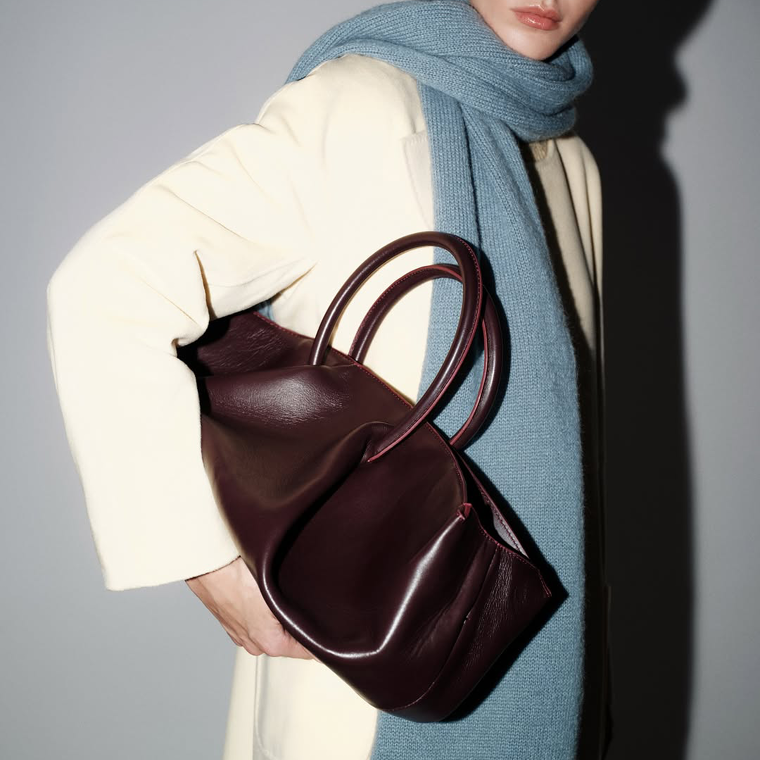 A person wearing a cream coat and a long, light blue knit scarf holds a large, slouchy burgundy leather tote bag with rounded top handles.